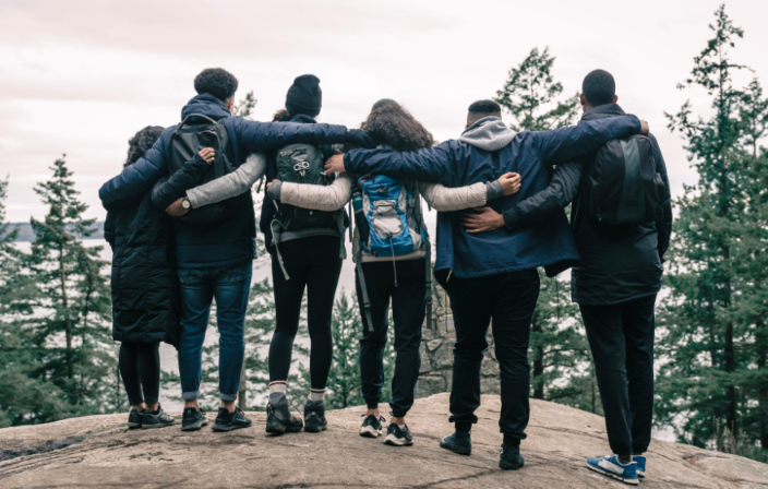 Eine Gruppe von Männern und Frauen stehen Schulter an Schulter umarmt an einer Klippe und schauen auf einen See.